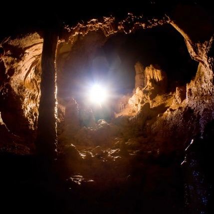 Вид изнутри узкого пещерного хода на яркий источник света у выхода; видны каменные столбы, влажный свод и контуры сталагмитов.