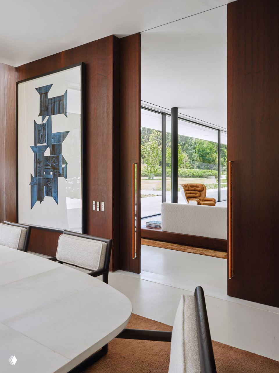 Проём между помещениями: крупная рамка с абстрактным постером, проход в гостиную с низкой мебелью и видом на зелёную зону за домом.