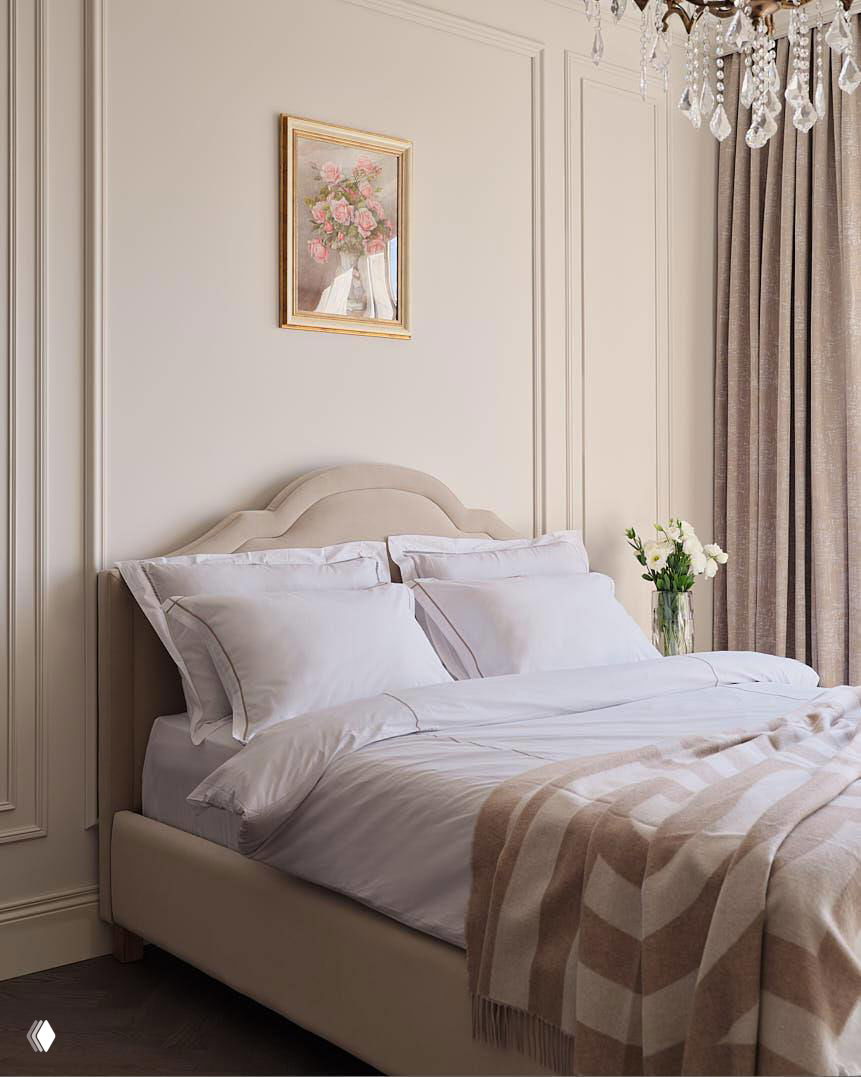 Спальня в мягкой бежевой гамме: изящное изголовье кровати, белое постельное бельё и плед в тон, рядом ваза с цветами на прикроватной тумбе.