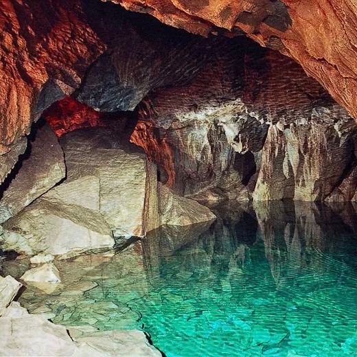 Подземное озеро с прозрачной бирюзовой водой, отражающей обломки известняка и тонкие сталагмиты на берегу в приглушённом свете пещеры.