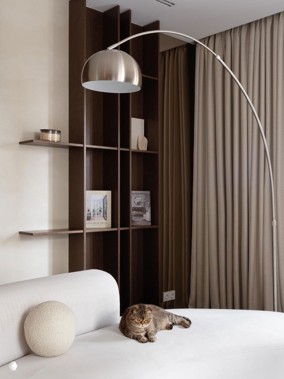 Светлая округлая софа у деревянной полки и торшера; на софе лежит пушистый коричневый кот, подчёркивающий домашнюю атмосферу интерьера.