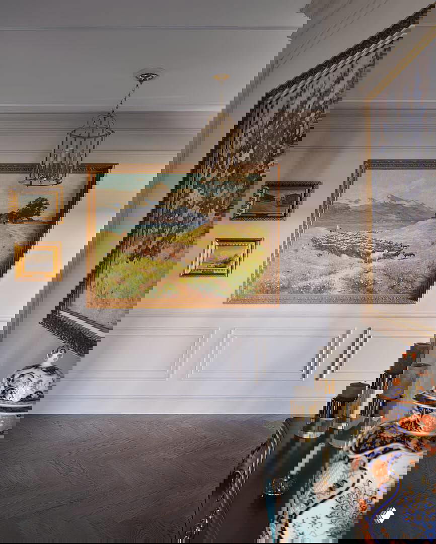 Прихожая с большими картинами в золочёных рамах, светлая стеновая панель и декоративная консоль с фарфоровым сосудом — акценты классического интерьера.