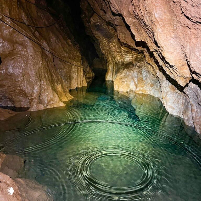 Узкий подводный коридор пещеры с прозрачной водой и зеркальной поверхностью, видны рябь от капель и тёмные своды над водой.