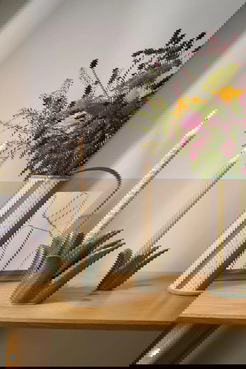 Декоративные предметы на деревянной поверхности: керамические подсвечники в форме цветка и высокая цветная ваза с полевыми цветами на фоне пастельной стены.