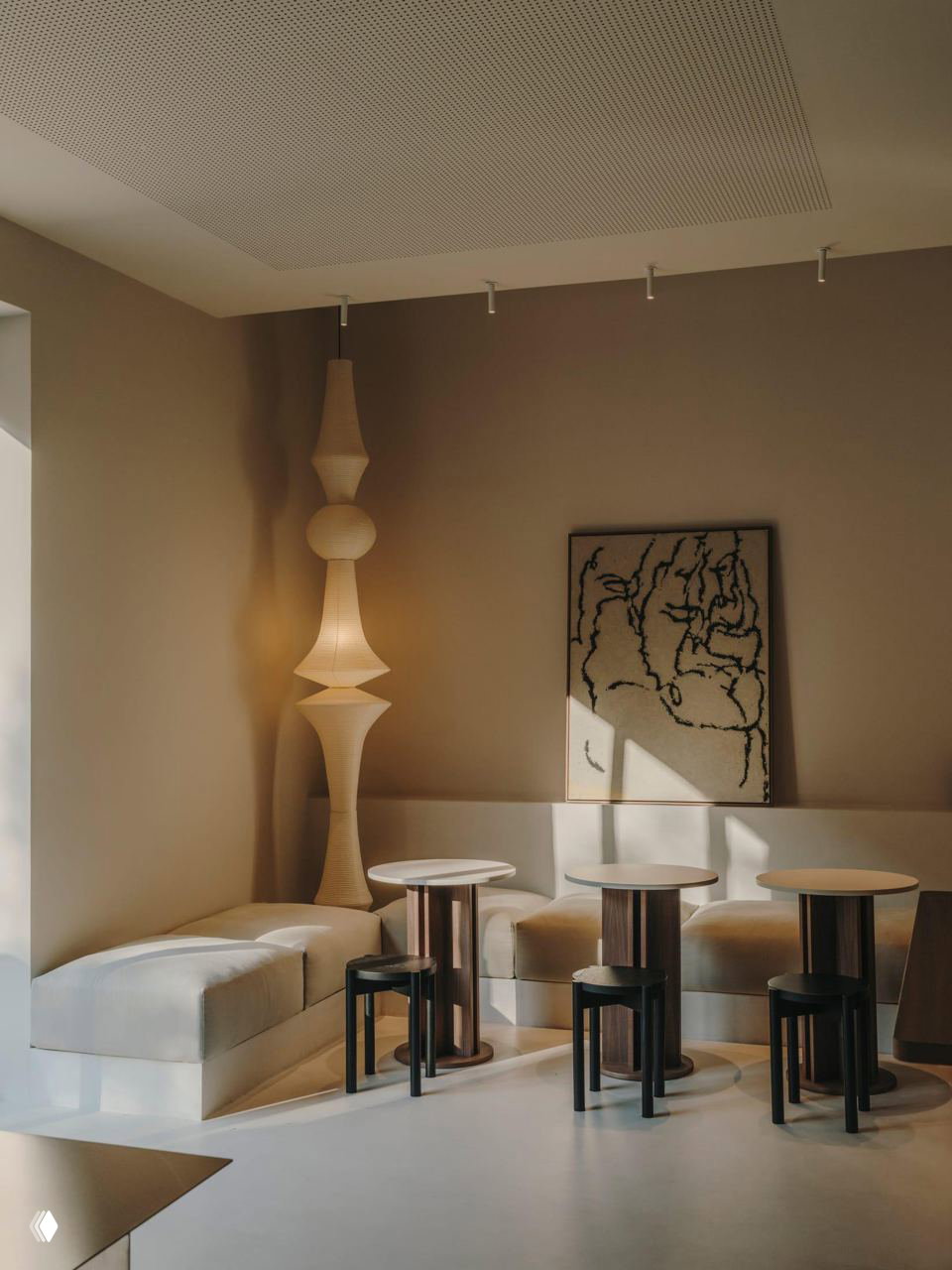 Уютный уголок кафе: низкие столы и мягкие пуфы у стены, авторская напольная лампа и картина на фоне нейтральных тонов интерьера.