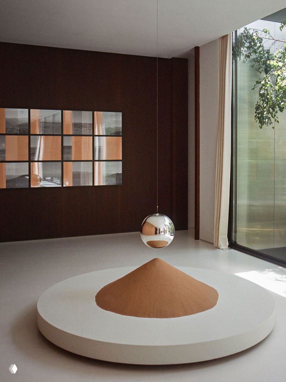 Минималистичная инсталляция: круглая подставка с насыпанной песочной формой и подвесной зеркальной лампой у большого окна с видом на сад.