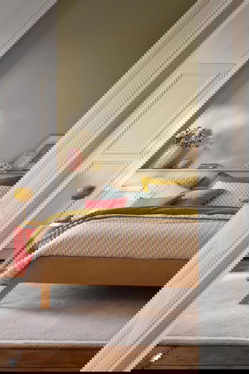 Спальня с деревянной кроватью, пастельным постельным комплектом, мягким ковром и акцентами оливковых и горчичных оттенков в спокойном загородном интерьере.