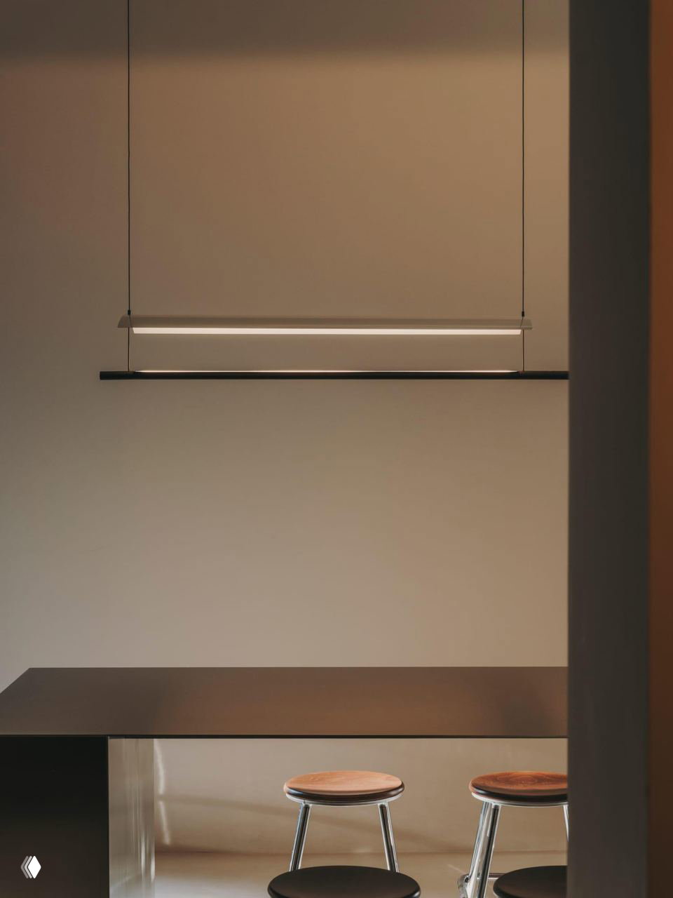 Минималистичная барная зона с подвесным светильником над столешницей и двумя высокими табуретами на фоне светлых, спокойных тонов помещения.