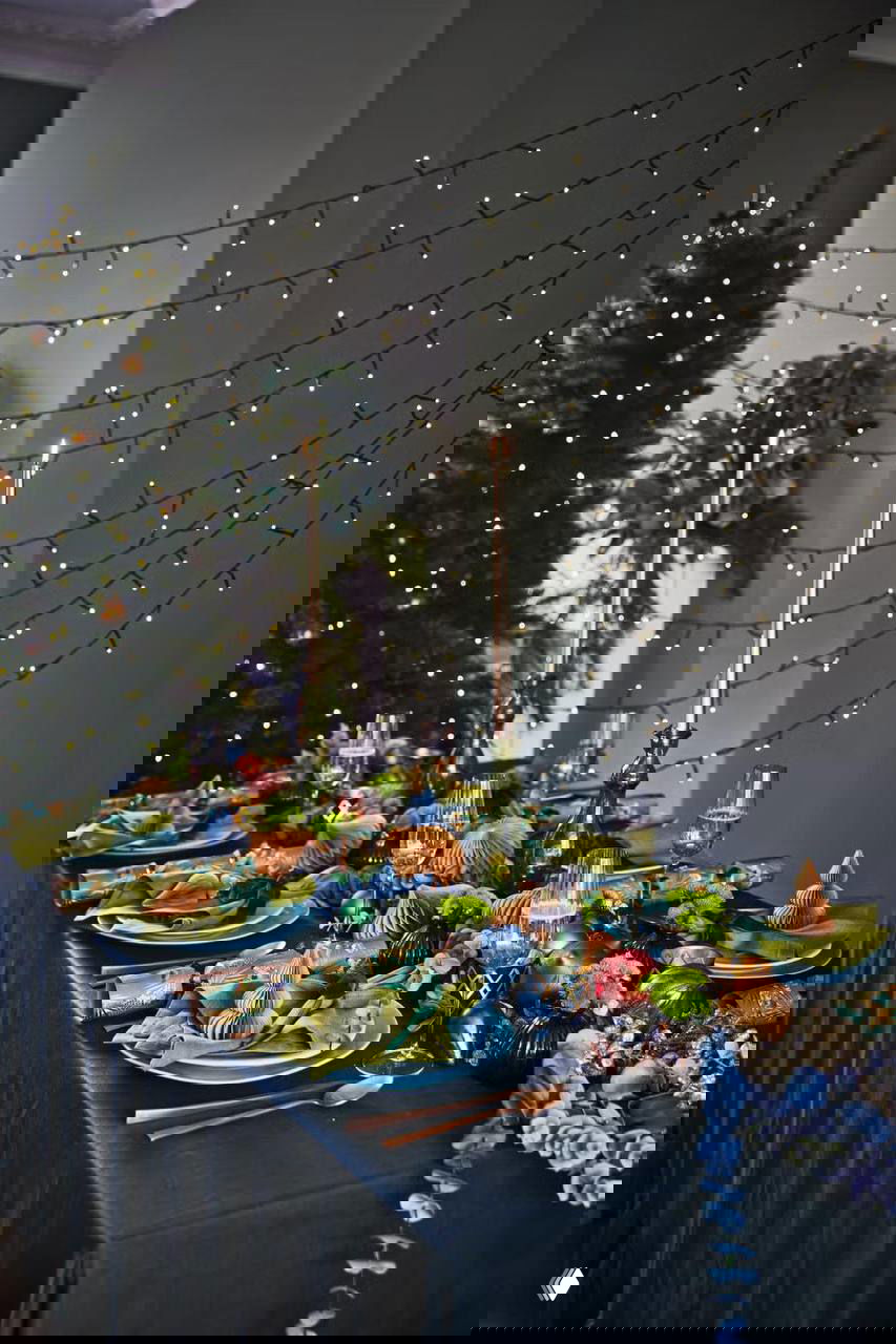 Элегантная праздничная сервировка с тёмной скатертью, зелёными и синими акцентами, стеклянной посудой и гирляндой световых огоньков на стене позади стола.