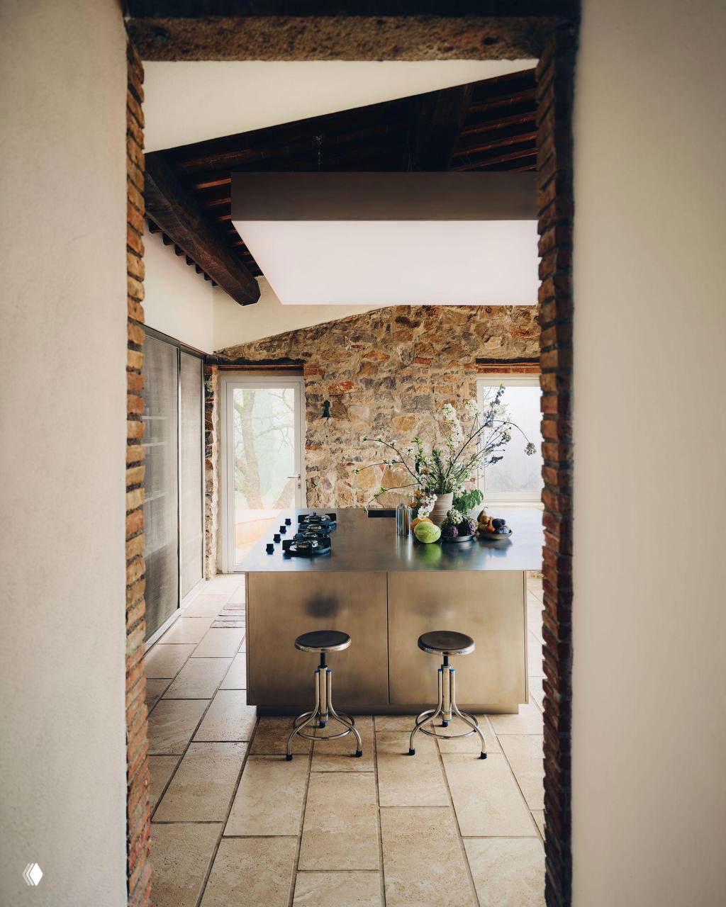 Вид на кухню с островом из хромированной стали, табуретами и каменной стеной Тосканы — сочетание традиции и индустриального хрома.