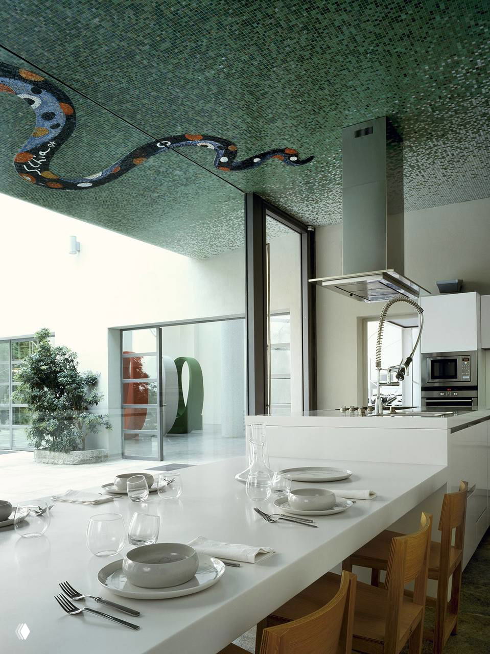 Белая кухня-столовая в доме Новембре: длинная белая столешница, стеклянная посуда и мозаичный зелёный потолок с изображением змеи.