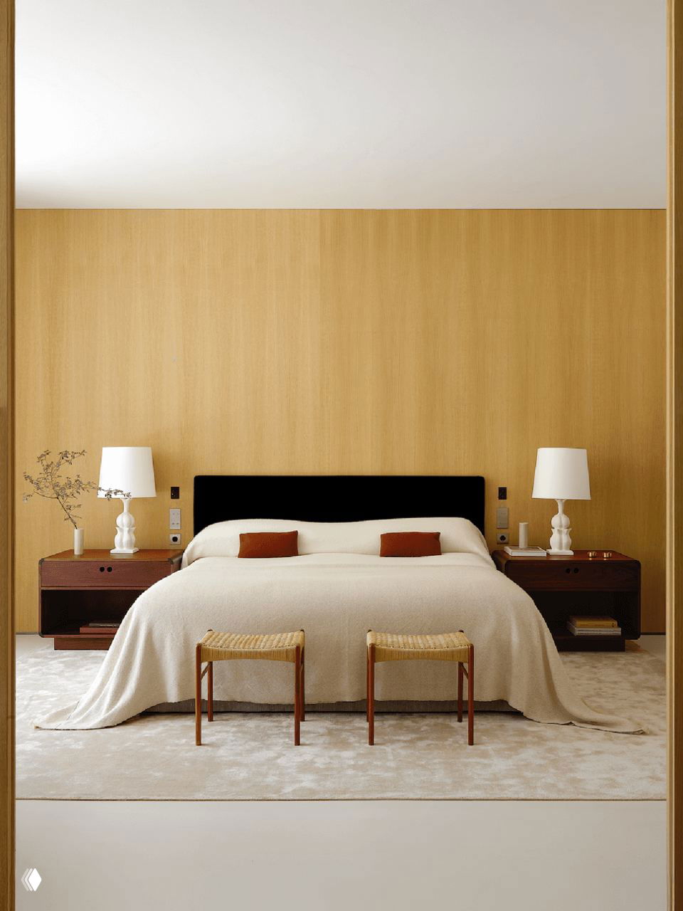 Спальня с тёплой деревянной панелью, большой кроватью в светлых тонах, тёмными прикроватными тумбами и ротанговыми табуретами у изножья.