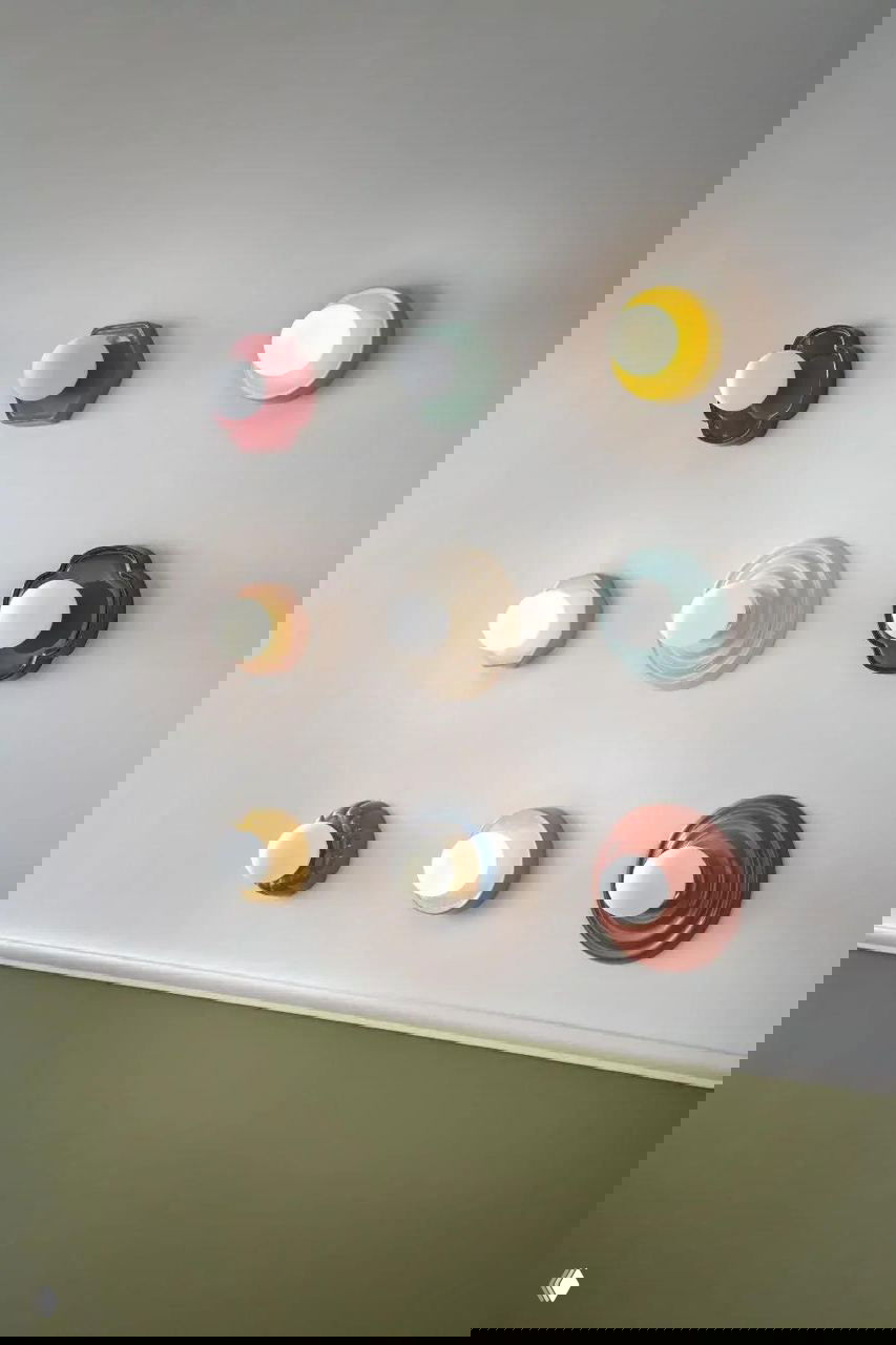 Настенные керамические светильники разной формы и цвета, расположенные в сетке на светлой стене — демонстрация керамического дизайна коллекции La Redoute.