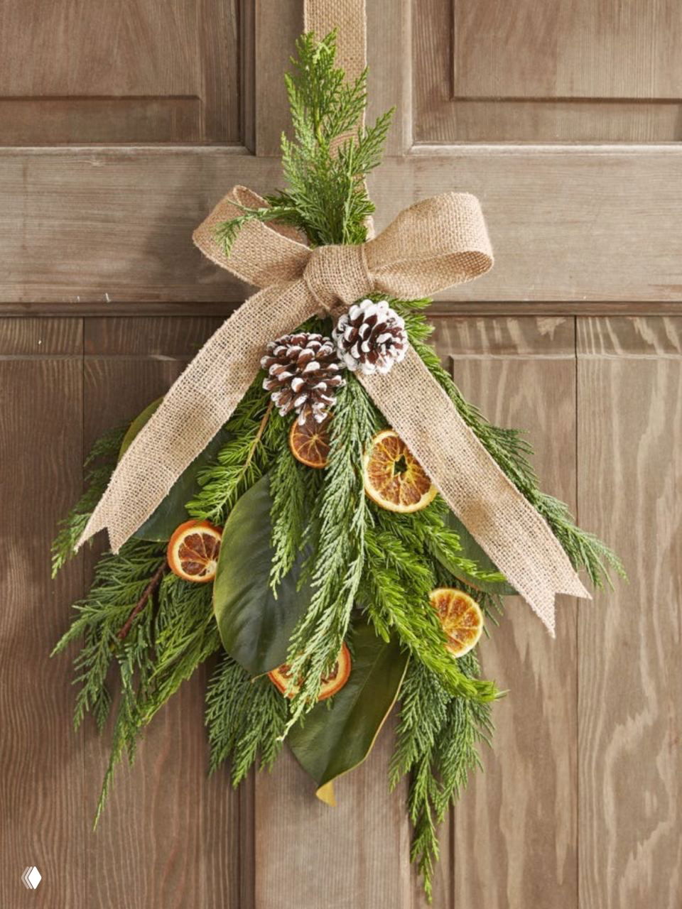 Дверной букет из веток хвои, шишек и сушёных ломтиков апельсина, перевязанный джутовой лентой — натуралистичный входной декор для праздника.