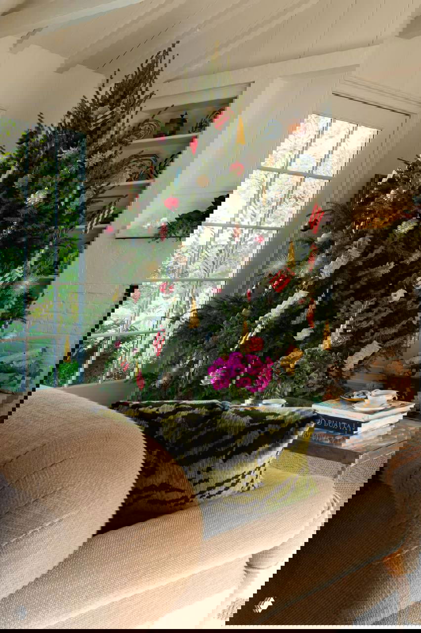 Большая рождественская ёлка у панорамного окна: традиционные игрушки и шары, рядом уютный диван и столик с книгами — тихий семейный уголок для праздника.