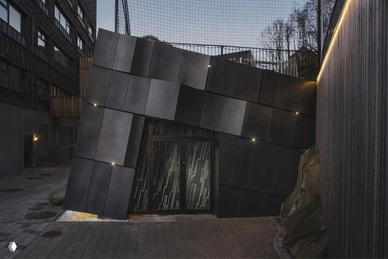 Ночной вид фасада: массив из ритмичных прямоугольных панелей, точечная подсветка у дверного проёма и деревянные ограждения на подходе к входу.
