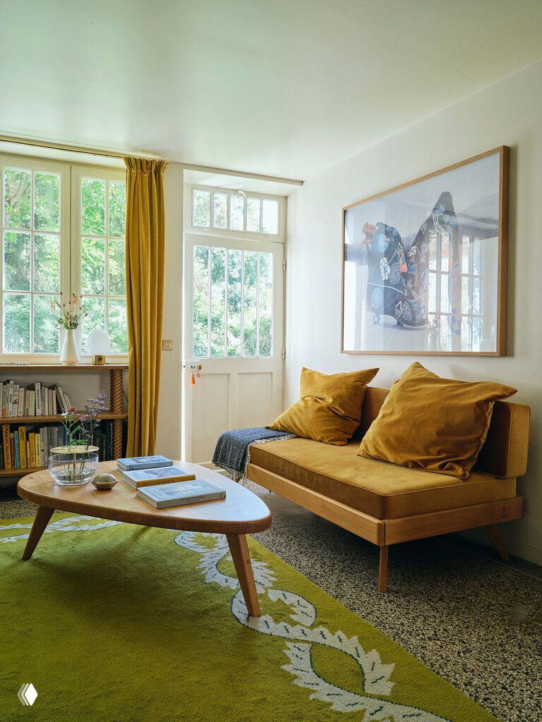 Гостиная у Сены с горчичным диваном, зелёным ковром и низким журнальным столом, большое окно открывает вид на сад.