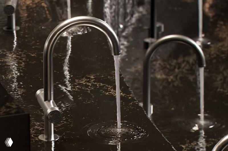 Крупный план смесителей Grohe Spa: металлические изливы и поток воды над тёмной мраморной столешницей с рябью и каплями