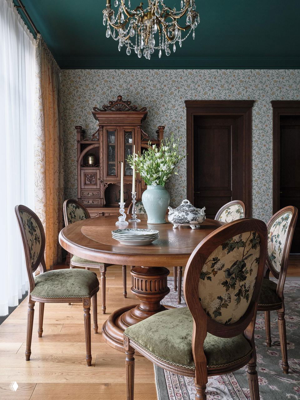 Столовая в английской классике: круглый деревянный стол, резные стулья с тканевой обивкой и деревянный буфет на фоне цветочных обоев.