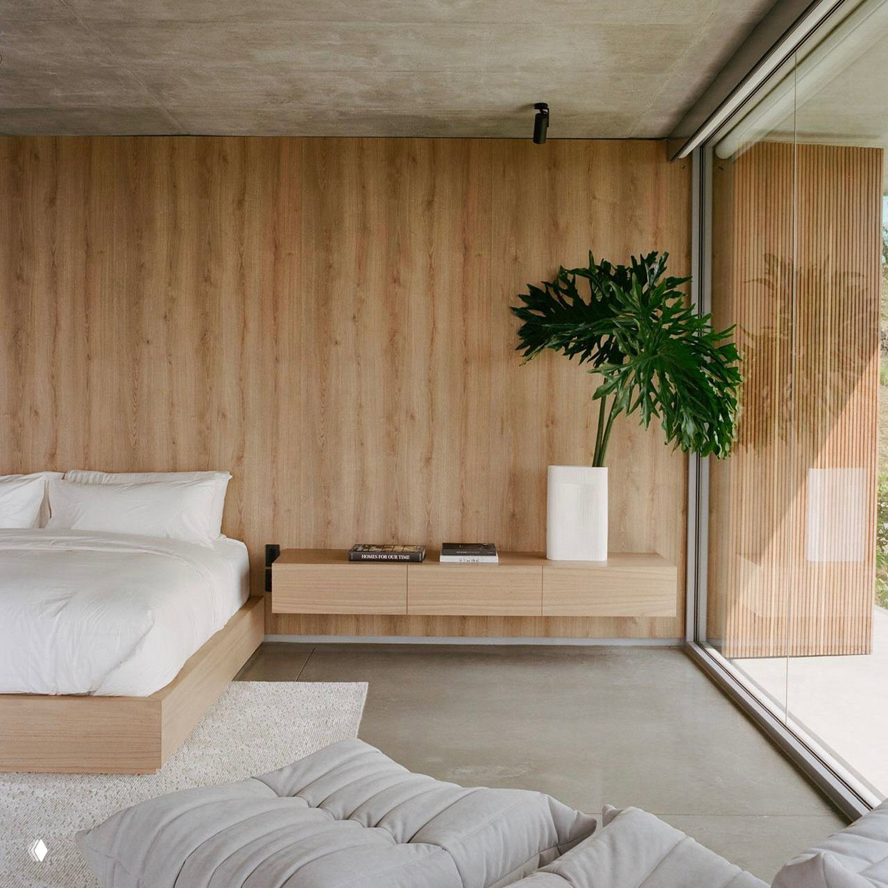 Минималистичная спальня с деревянной стеной, низкой кроватью, бетонным полом и большим панорамным окном с видом на зелень