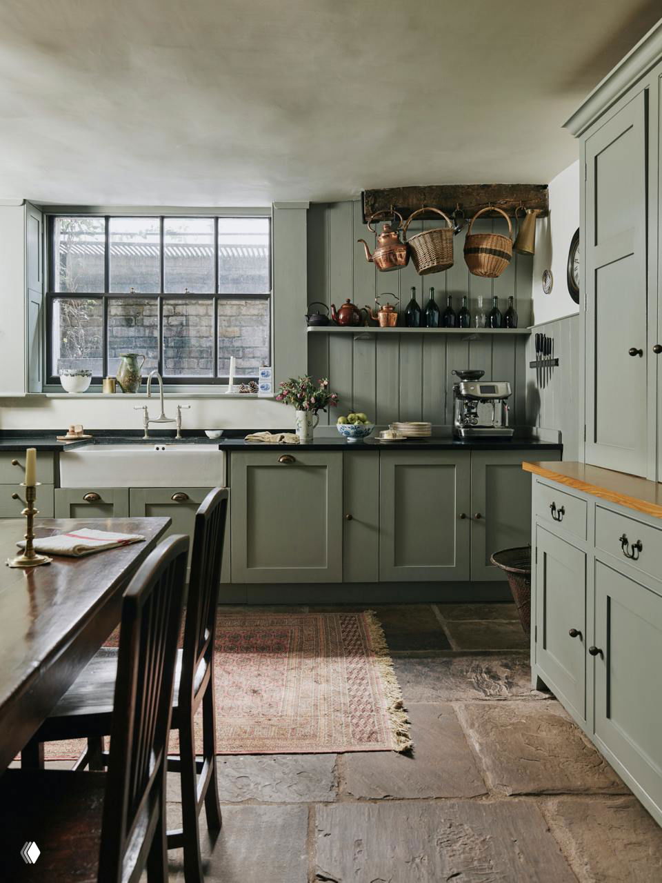 Кухня в лондонском доме: фасады мягкого зелёного цвета, открытые полки с медной утварью, широкий оконный проём над мойкой и массивная столешница на фоне каменного пола.