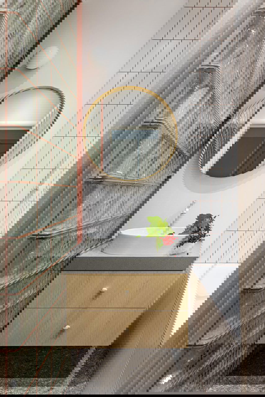 Ванная комната с массивной стеклоблоковой перегородкой, круглым зеркалом и компактной тумбой‑раковиной; сочетание текстур плитки и мрамора в небольшом пространстве.