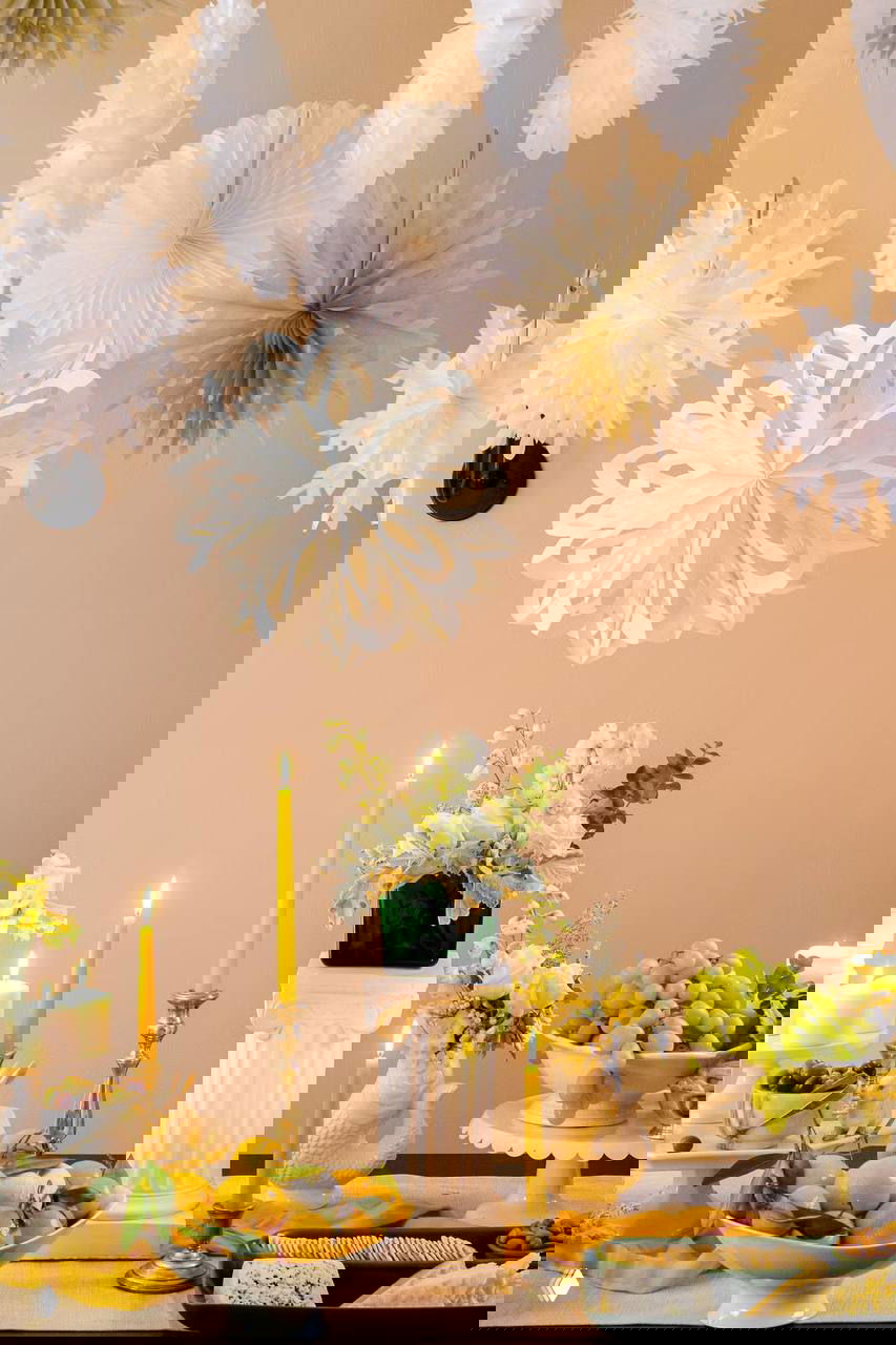 Нежный праздничный стол с висящими бумажными снежинками над ним, свечами и светлыми фруктовыми композициями — лёгкий и воздушный декор для торжества.