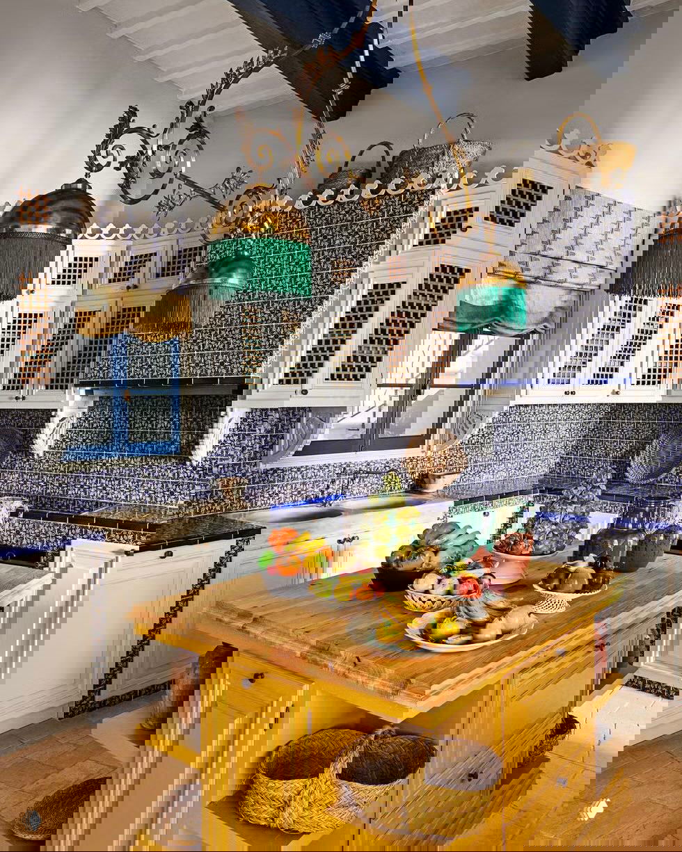 Яркая кухня в средиземноморском стиле с жёлтым деревянным островом, фруктами на столешнице и декоративной настенной плиткой вокруг.