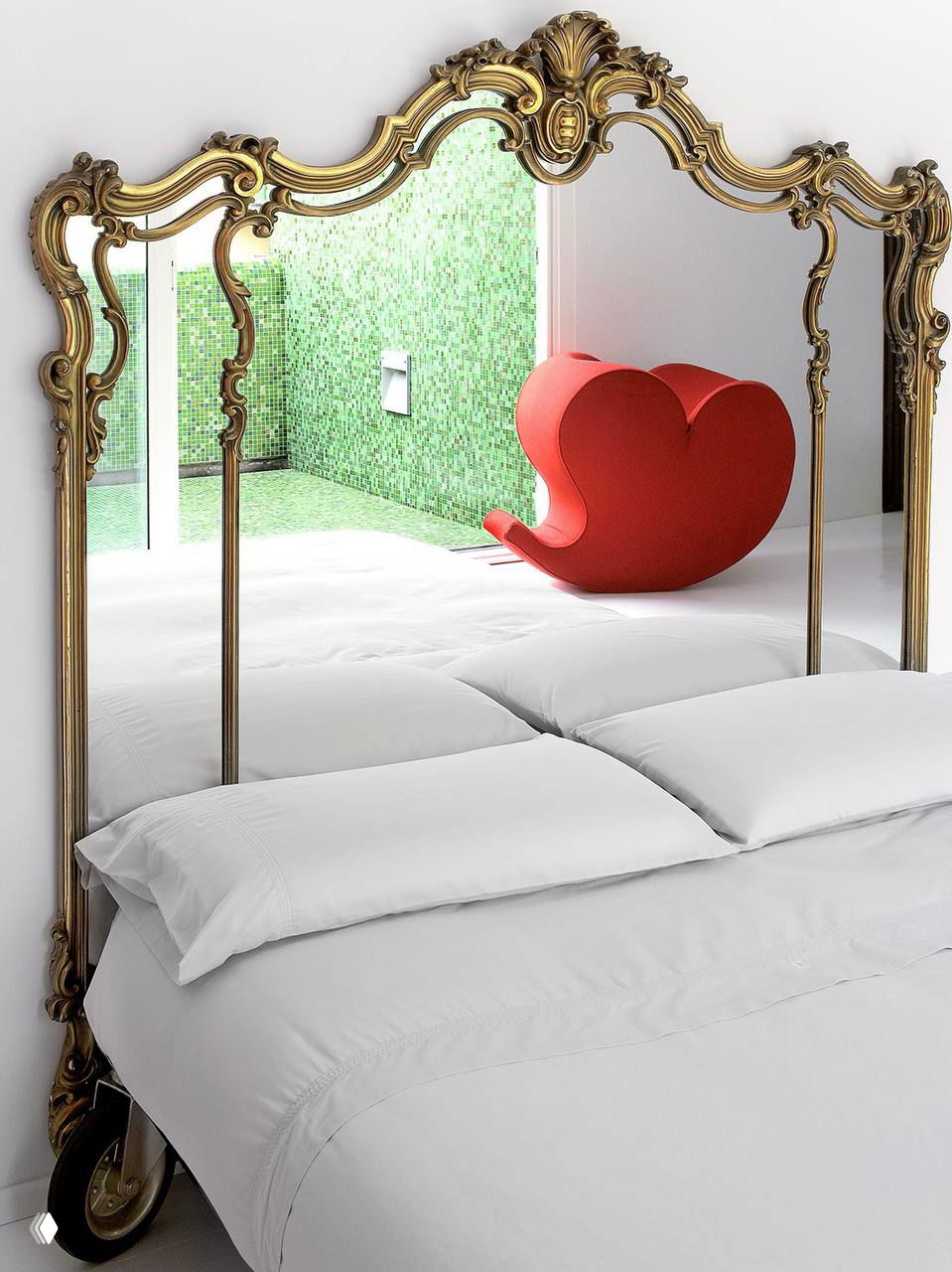Спальня с большим резным зеркалом в золочёной раме, белое ложе и яркий красный дизайнерский кресло на заднем плане.