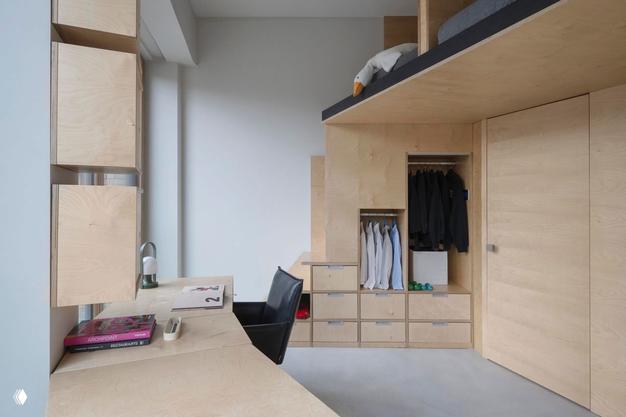 Детская/рабочая зона с модульной мебелью из светлой фанеры: встроенные шкафы, рабочая поверхность и высокий спальный подиум над гардеробом.