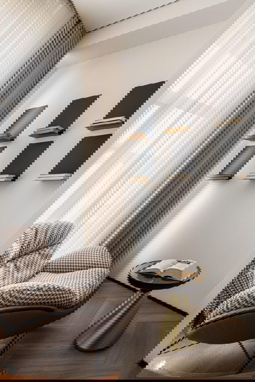 Уютный уголок с креслом и тонкими полками для книг у высокого окна, лёгкие шторы пропускают дневной свет — спокойная читальная зона.