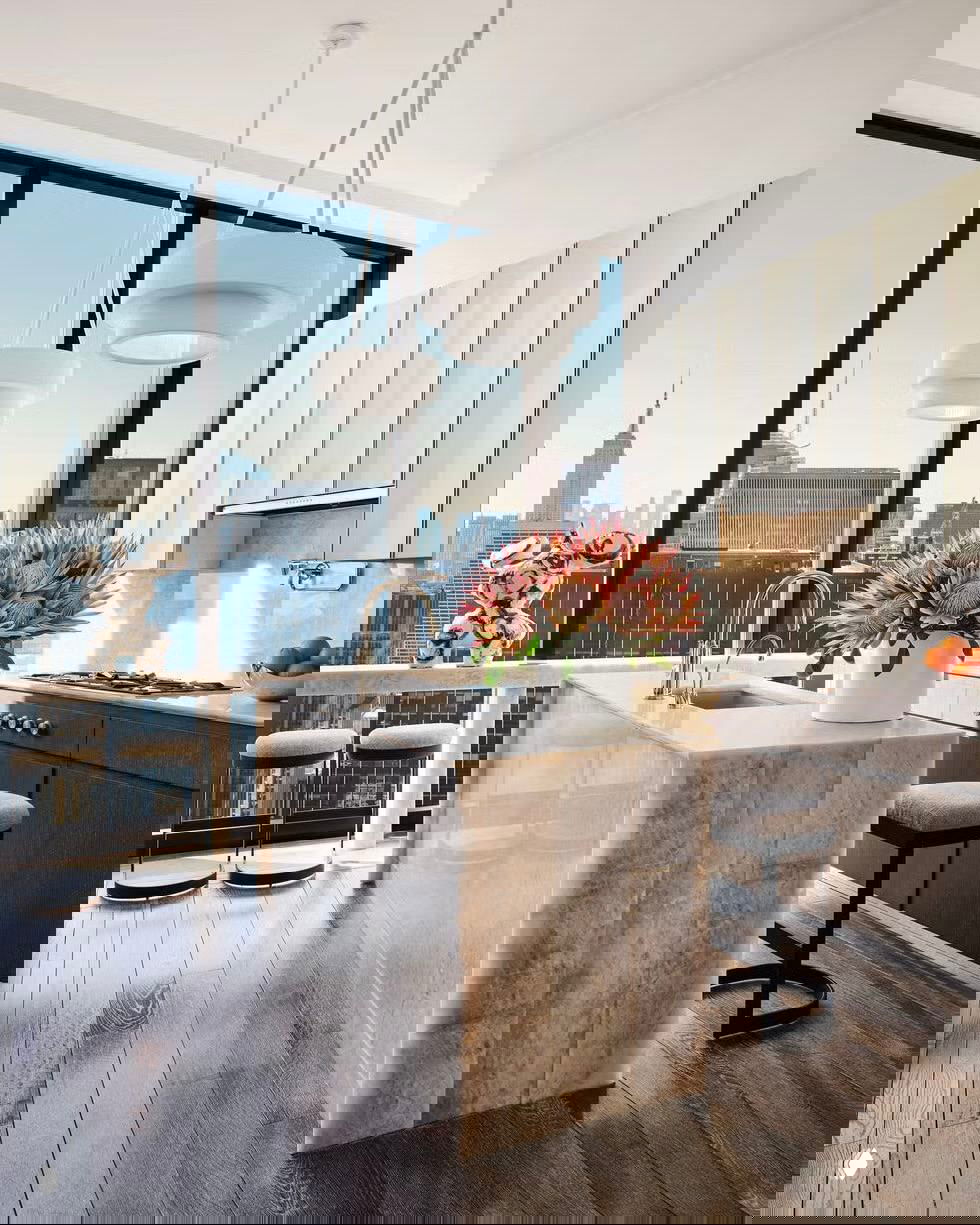 Современная городская кухня с высокими окнами, минималистичным мраморным островом, мойкой в острове и видом на городской пейзаж.