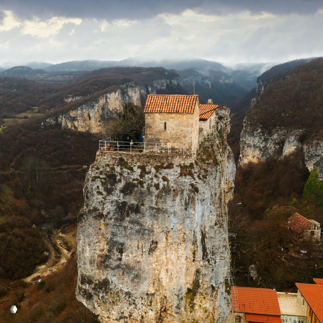 Крупный план столпа Кацхи: каменная капелла с черепичной крышей на узкой вершине скалы и временными лестницами, ведущими вверх.