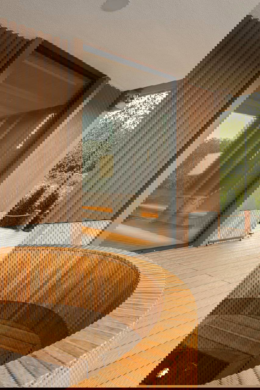 Наружная терраса с круглой деревянной купелью и входом в парную за стеклянной дверью; обеденная зона у воды и отделка композитной террасной доской.