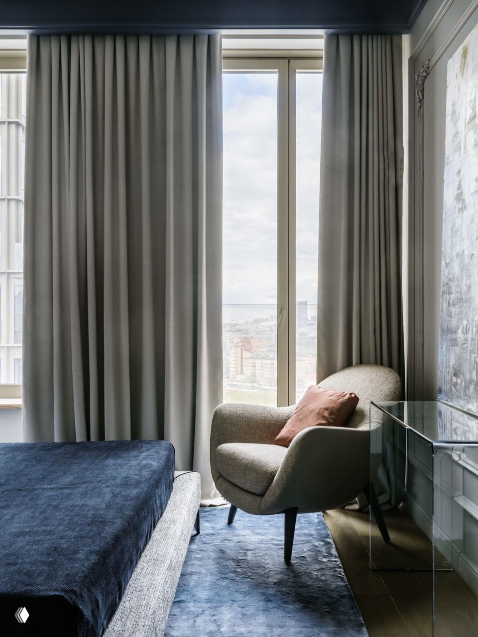 Спальня с креслом у окна: синий текстиль на кровати, мягкое кресло и полупрозрачные шторы, спокойная палитра и акцентная подушка терракотового оттенка.