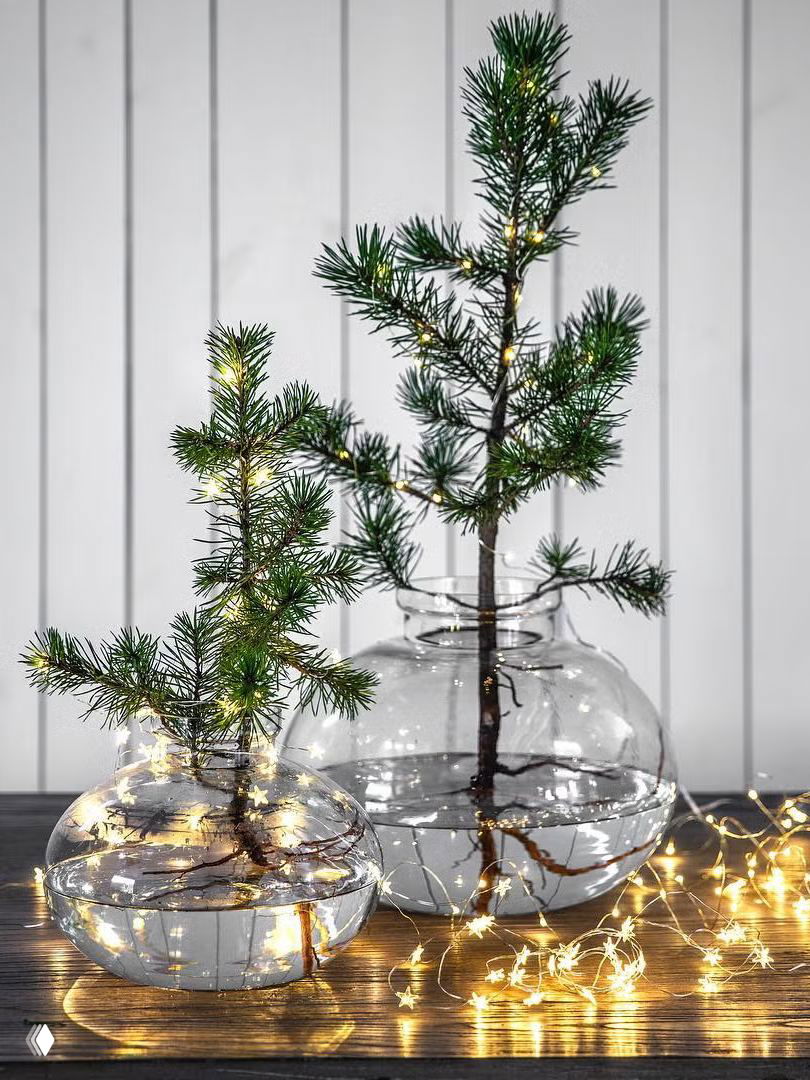 Небольшие веточки сосны в прозрачных круглых вазах с водой и тонкой гирляндой — скандинавская и простая идея праздничного светового декора.