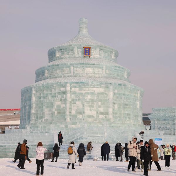 Куполообразная массивная ледяная постройка и толпа посетителей у подножия, плотная кладка блоков льда и яркие зимние одежды людей.