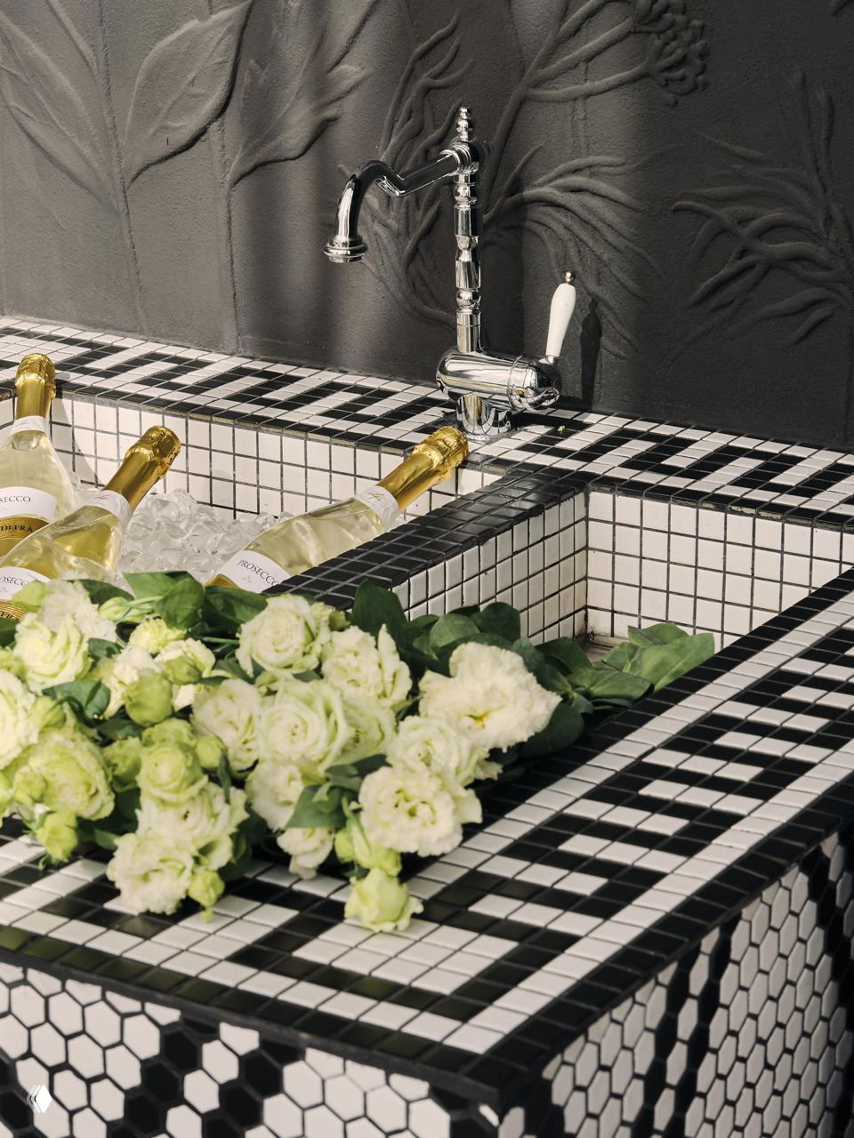 Деталь интерьера: мозаичная чаша-холодильник для шампанского с белыми цветами на переднем плане, декоративная плитка в чёрно‑белой гамме.