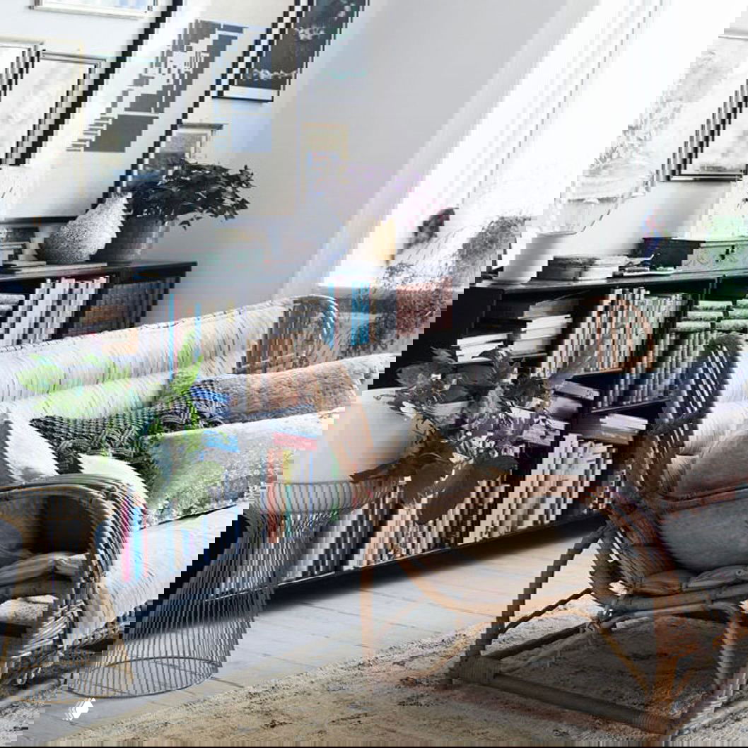 Библиотечный уголок: плетёный диван с множеством подушек на фоне низкой полки с книгами и комнатными растениями, светлый напольный рисунок и текстуры для уюта