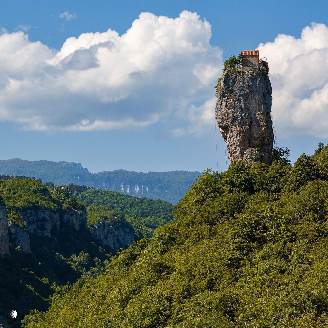 Дальний вид на тонкую колонну-скалу с церковью на вершине, столп Кацхи на фоне обширных горных хребтов и зелёных лесов.