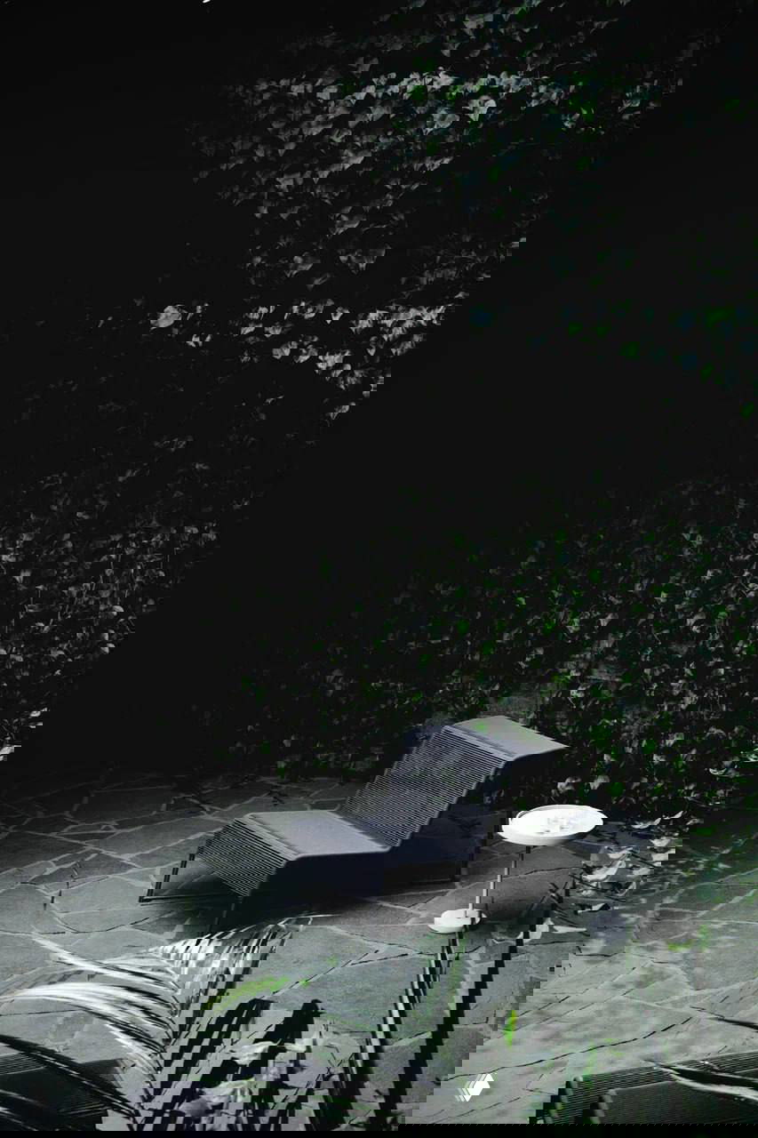 Лаунж-кресло на тёмном фоне у стены из плюща, каменная плитка под ногами и небольшой белый приставной столик в кадре