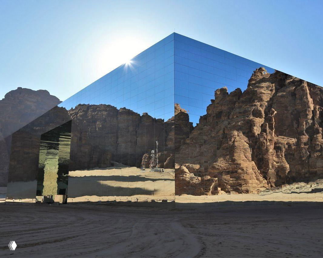 Зеркальный куб‑павильон в пустынном пейзаже: фасады отражают окружающие скальные образования и яркое небо, создавая иллюзию слияния с ландшафтом.