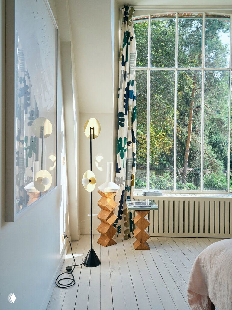 Уютный угол спальни с высоким оконным проёмом, узорными шторами, оригинальными деревянными табуретами и напольным торшером.