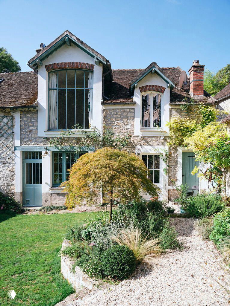 Фасад дома на берегу Сены с каменной кладкой, большими окнами и ухоженным садом перед домом с гравийной дорожкой.