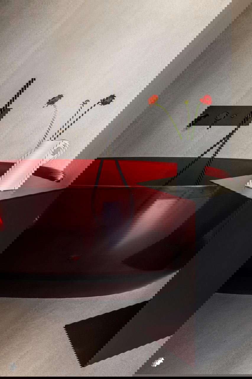 Ванная с прозрачной тёмно‑красной чашей ванны, чёрным смесителем и белой керамической кувшинной вазой с цветами — выразительный акцент в интерьере.