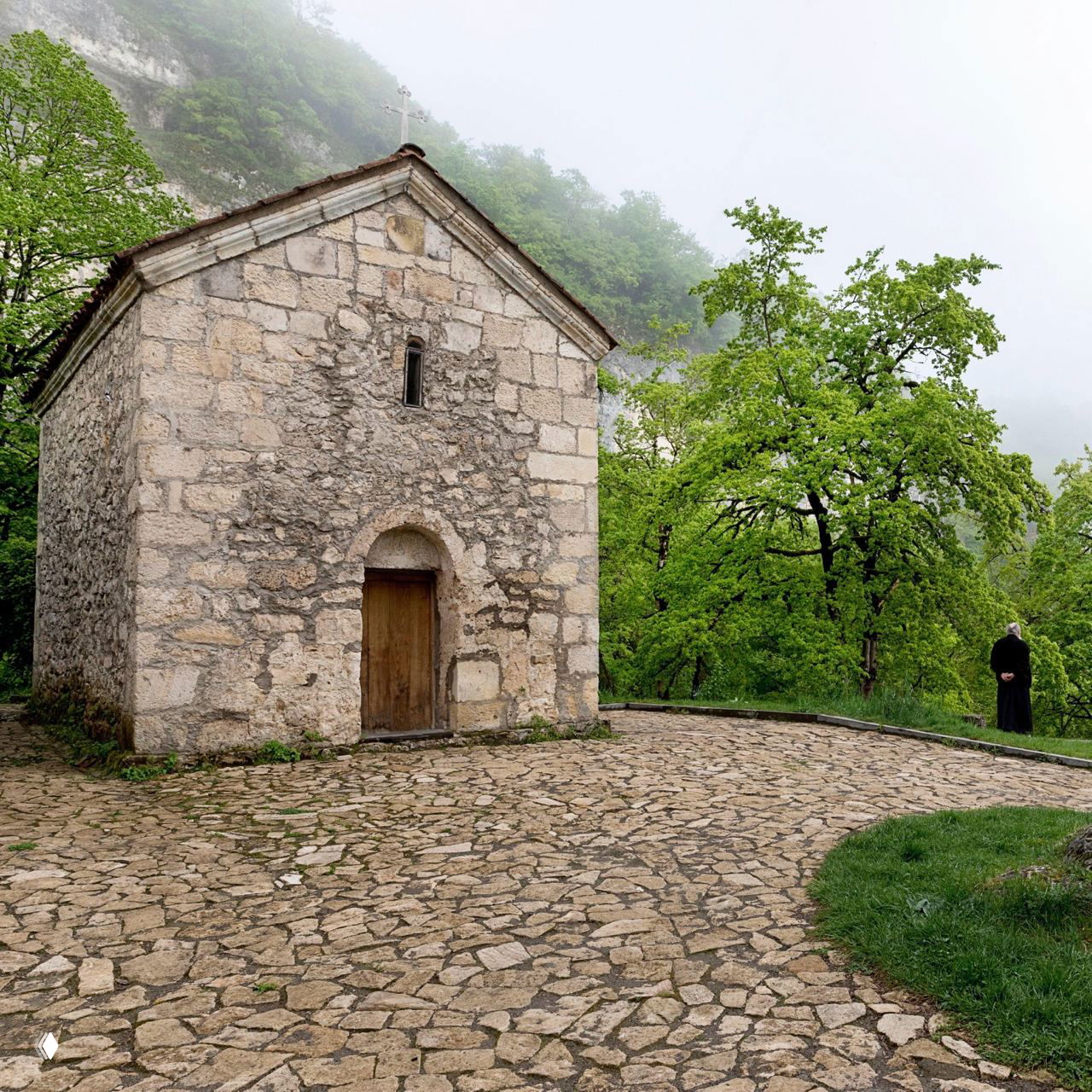 Малый каменный храм и мощёная площадка у подножия столпа Кацхи: булыжная дорожка, зелёные деревья и монах на краю снимка.