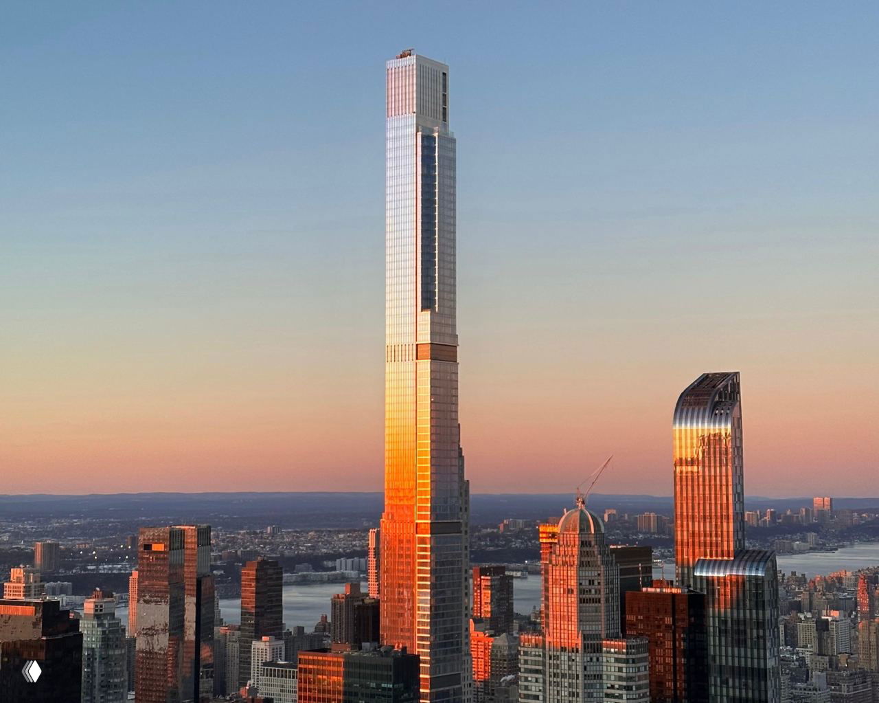 Высокий стеклянный небоскрёб на закате, фасады которого отражают тёплые оттенки неба и окружающих зданий в городской деловой зоне.