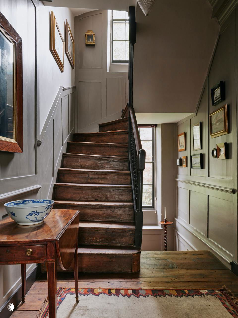 Лестничный пролет с потёртыми деревянными ступенями и высоким оконным проёмом: панелированные стены, картины в рамках и тёплый рассеянный свет из окон пролёта.