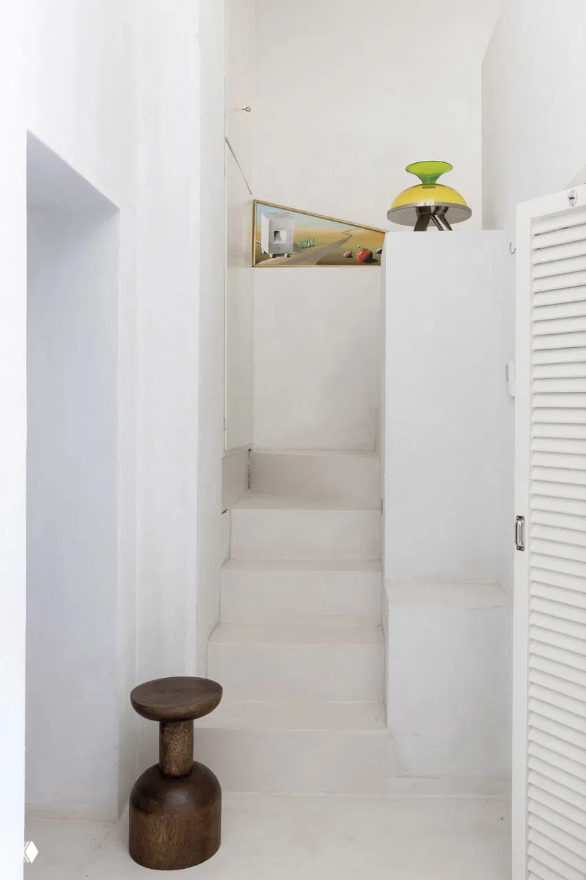 Узкая белая лестница и ниша в компактном доме: деревянный низкий табурет и декоративный элемент на фоне чистых стен и строгих форм.