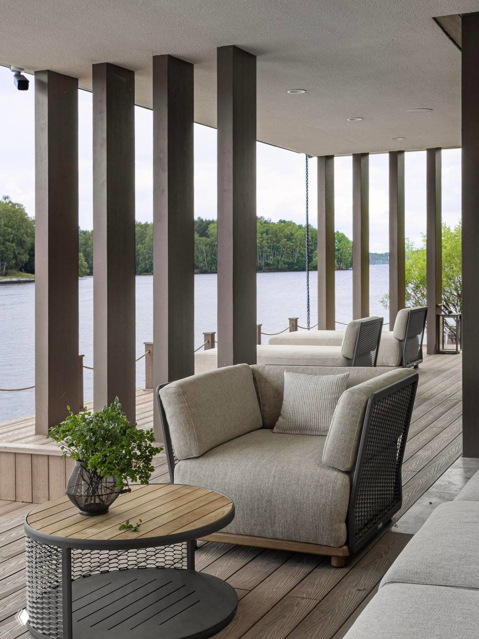 Открытая летняя зона у воды с креслами и столиком на палубе; колонны и ограждение подчёркивают связь внутреннего пространства с рекой и природой.